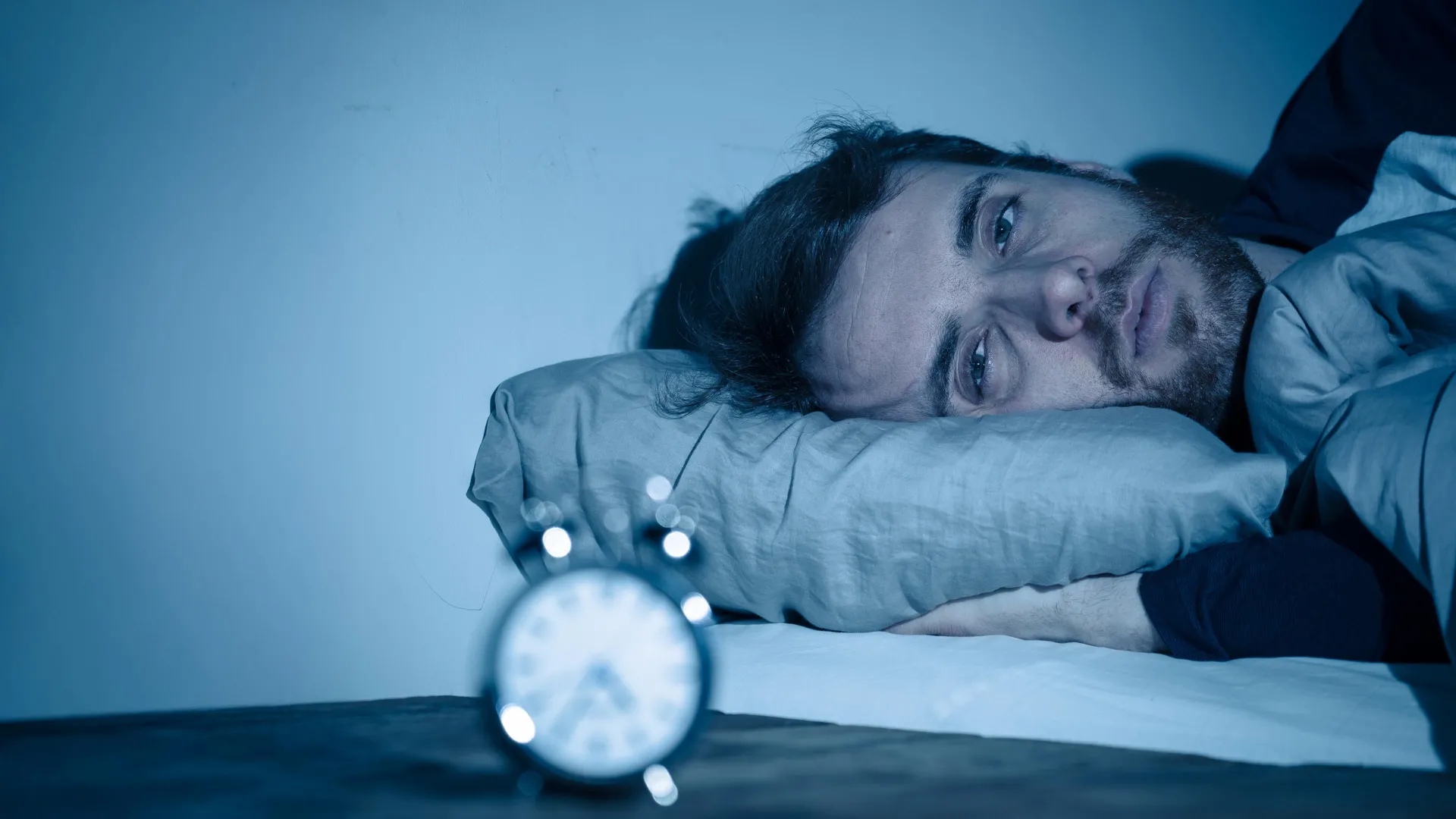 A man lies awake in bed at night, checking the time on his alarm clock, illustrating sleep deprivation and insomnia